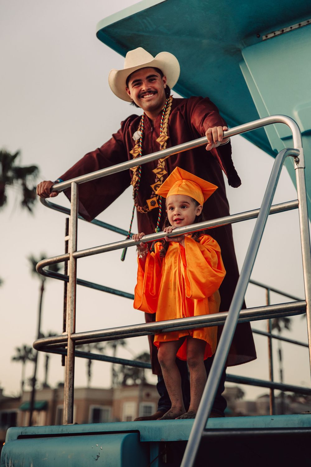 Man in cowboy hat and maroon graduation gown stands with child in orange cap and gown on lifeguard tower.