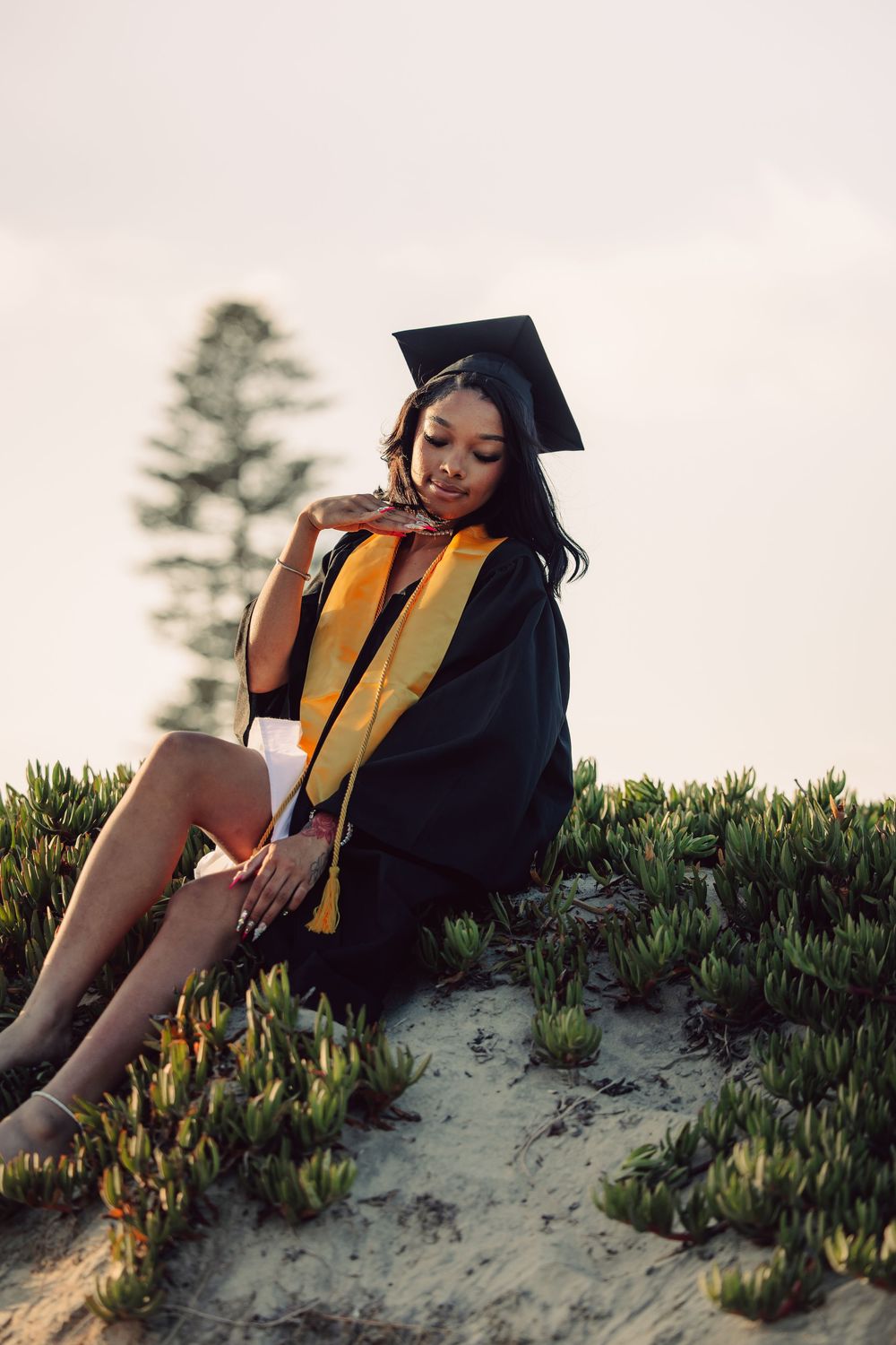 Young woman in black graduation gown and cap with gold stole sits on sandy hillside among green plants.