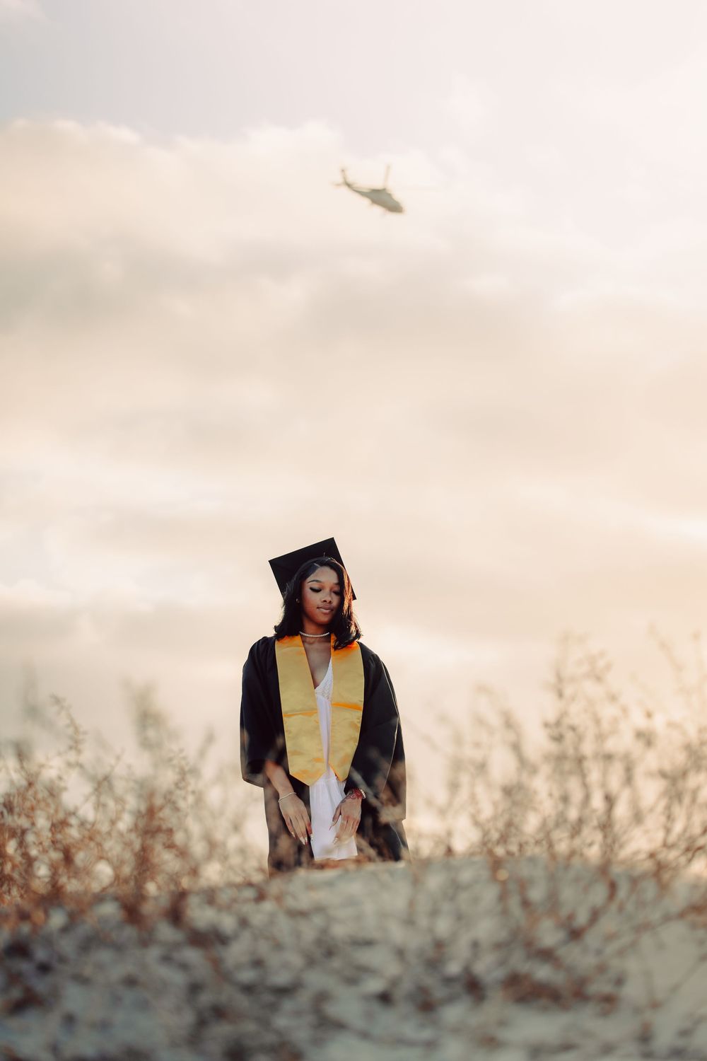 A young woman in graduation cap and gown stands outdoors at sunset with a helicopter visible in the cloudy sky.