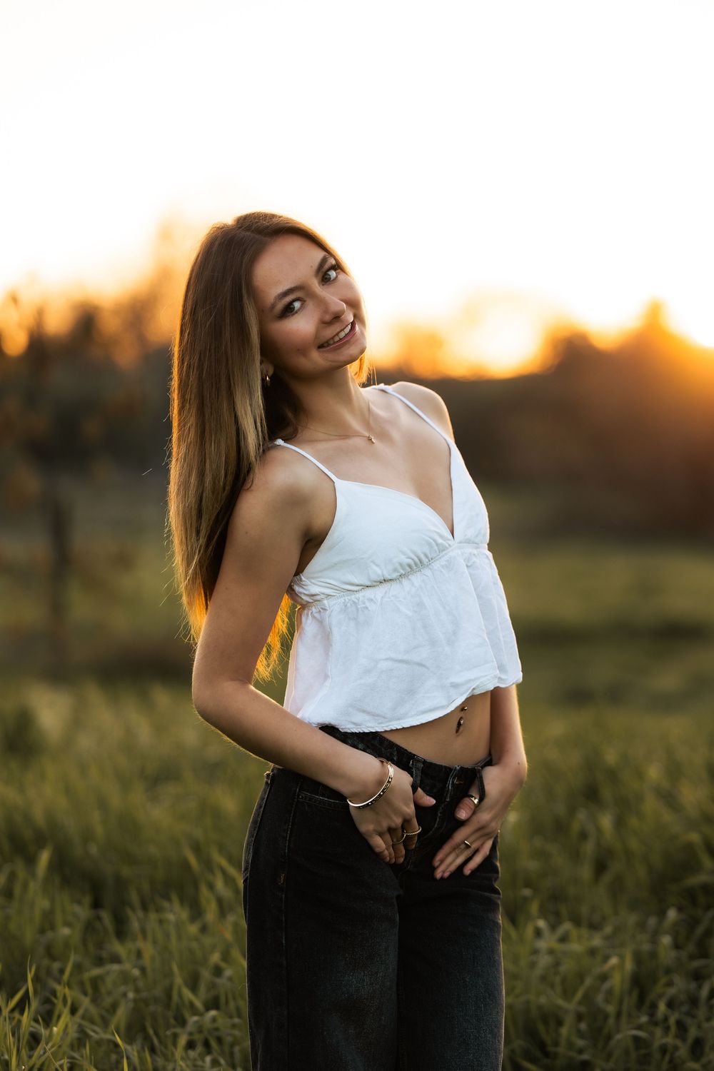 Young woman in white crop top and jeans smiling outdoors during golden hour sunset in a green field.
