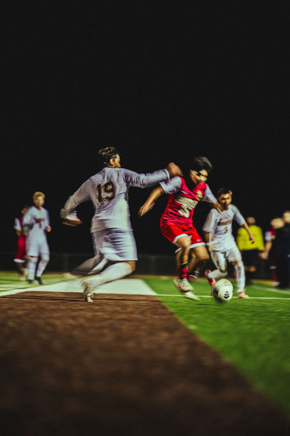 Soccer players in white and red uniforms compete for the ball during a nighttime match on an illuminated field.