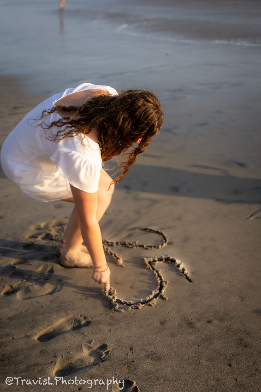 Person in white dress drawing a heart shape in wet sand on a beach at sunset.