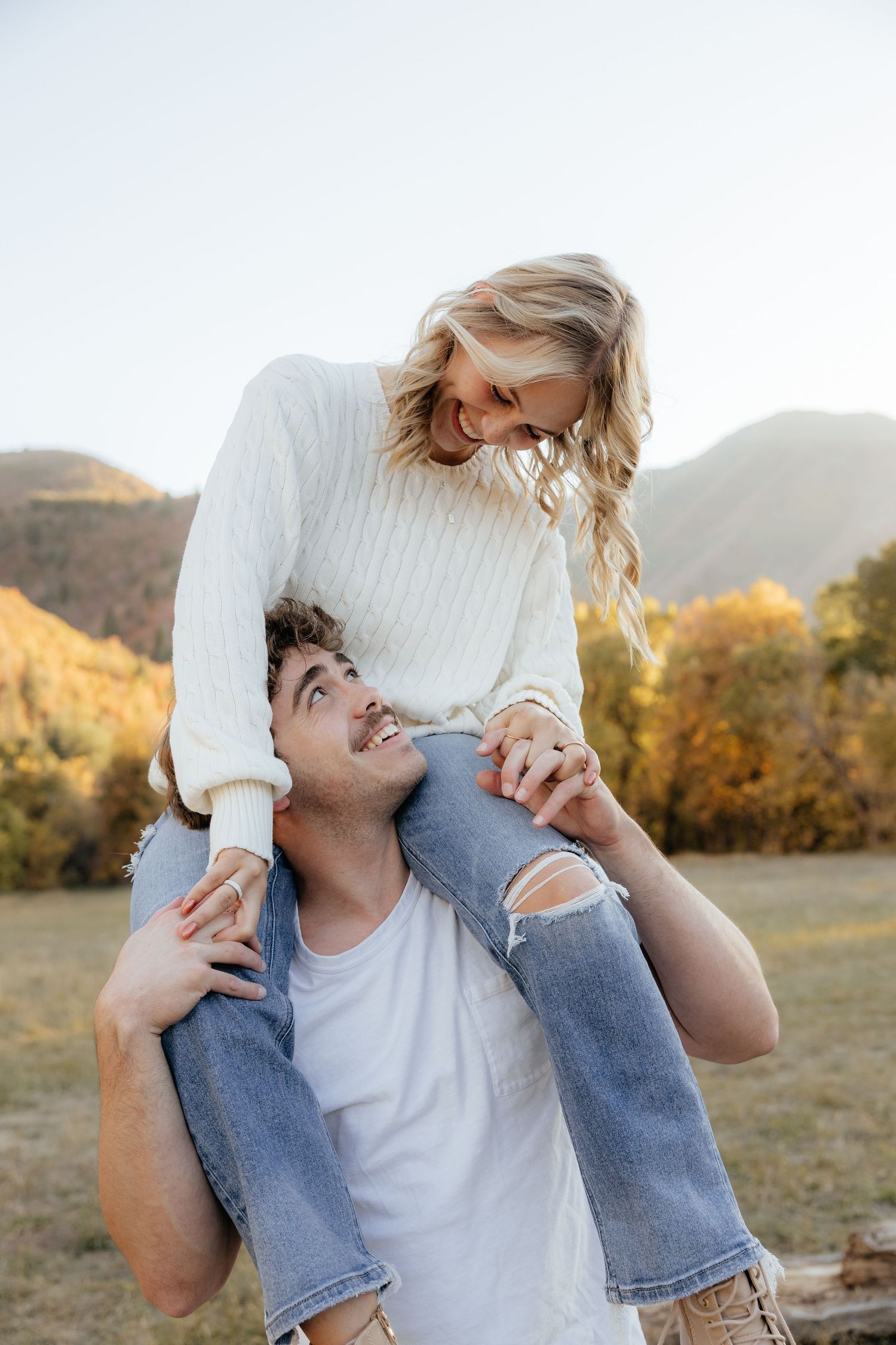 Couple laughing outdoors in autumn mountains, woman sitting on mans shoulders in white sweater.