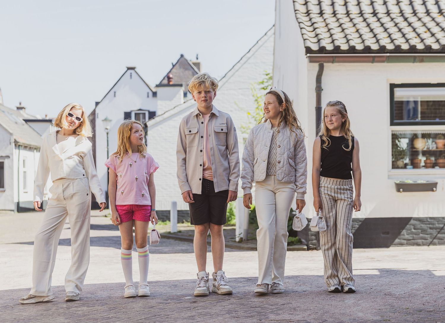 Five people of different heights standing in a row outside white buildings on a sunny day. Kinderkleding promotie fotografie