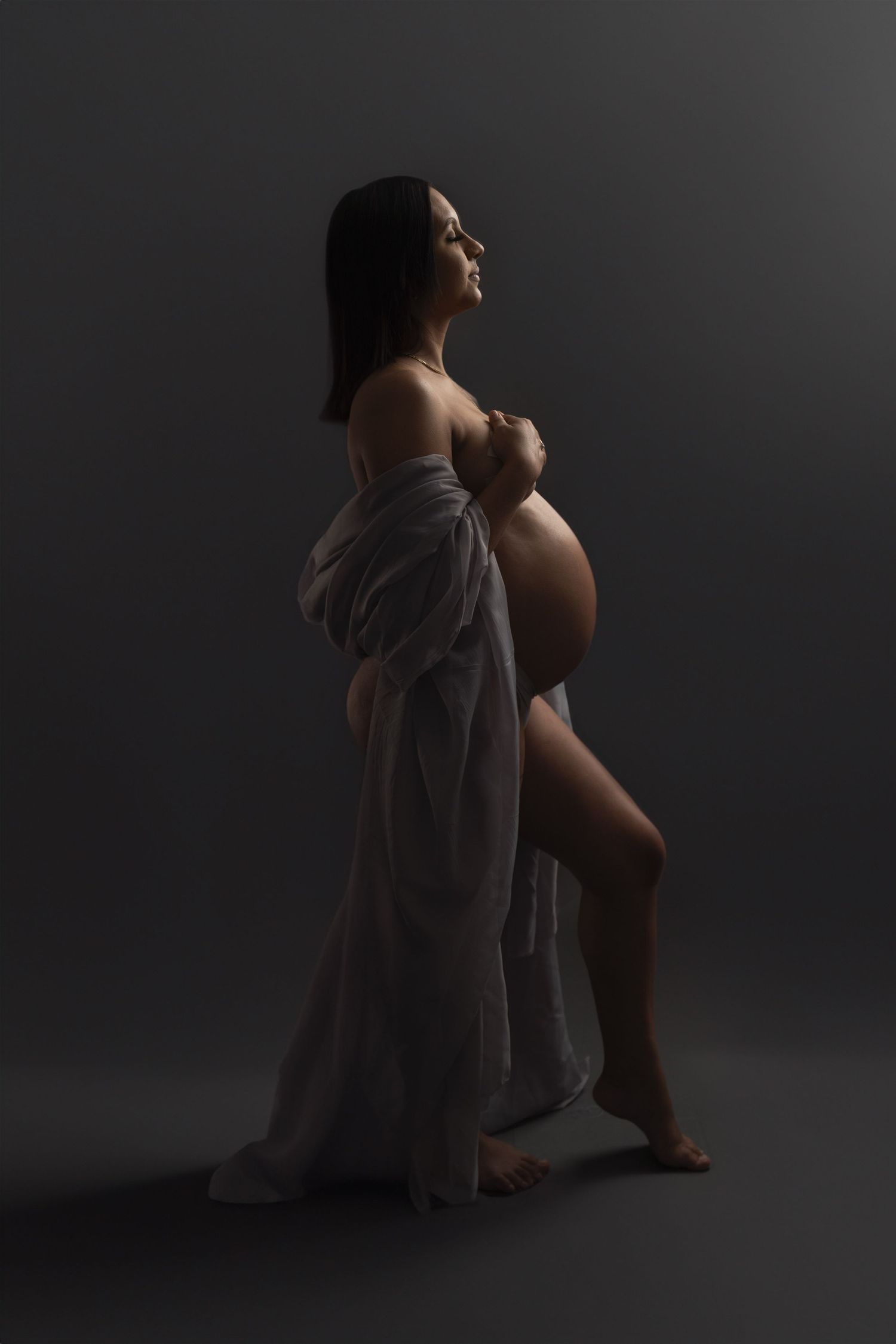 Dramatic maternity portrait with flowing fabric against dark background in artistic lighting.