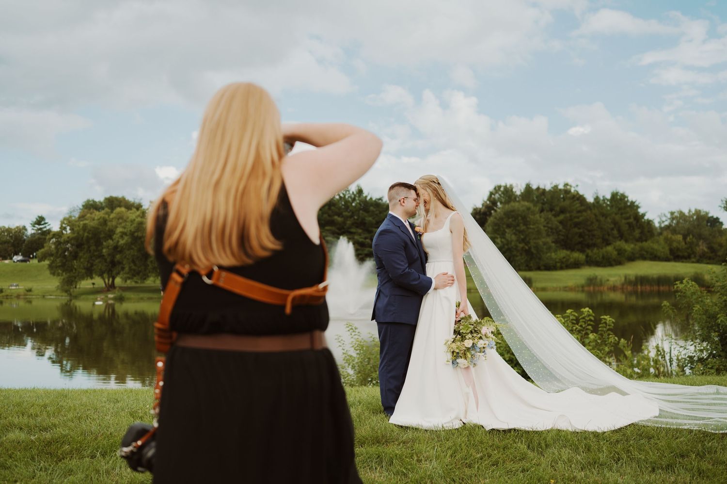 Wedding photographer captures couple posing by a scenic lake with flowing veil and lush green landscape.