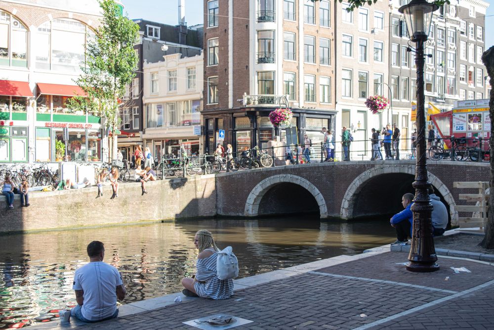 Canal bridge in Amsterdam with locals relaxing along the waterfront on a sunny day.