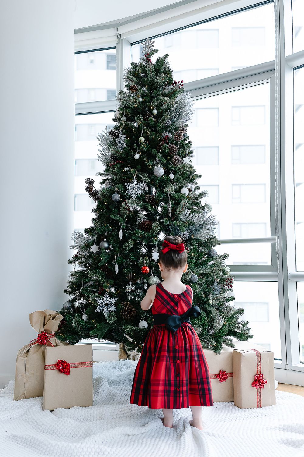 A child in a red plaid holiday dress admires a tall Christmas tree with wrapped presents beneath it.