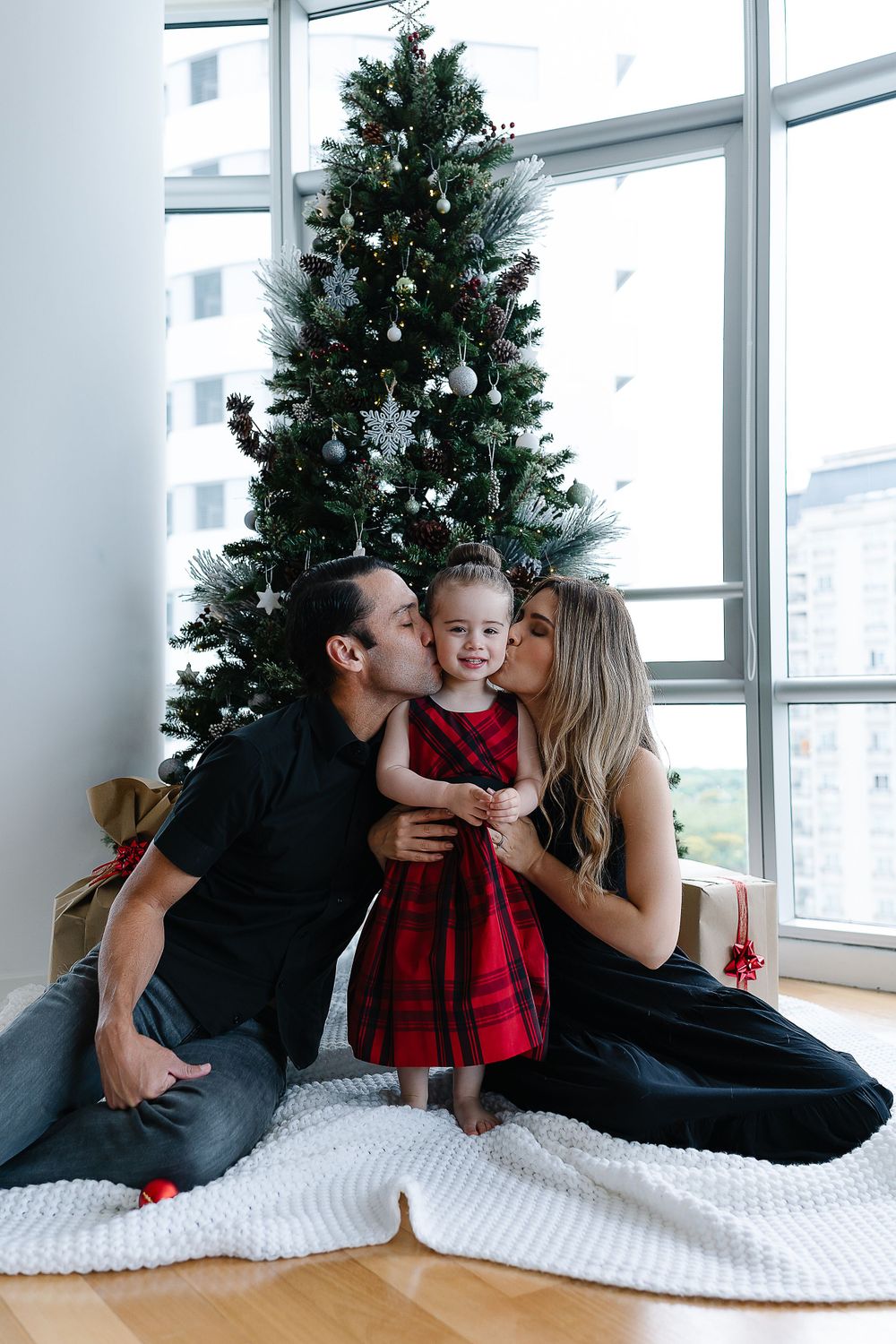 A festive family moment in front of a decorated Christmas tree with a child wearing a red plaid dress.
