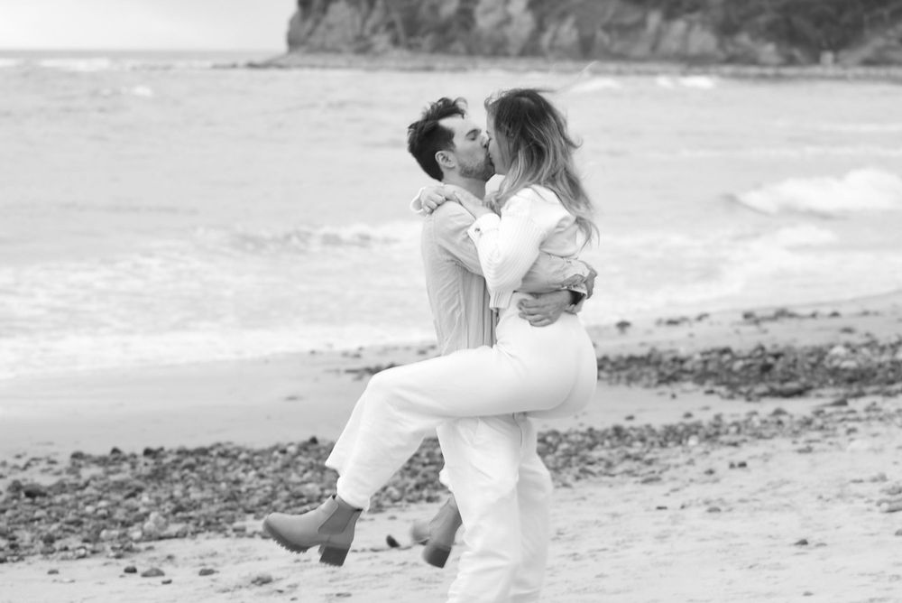 A romantic couple sharing intimate moments on a rocky beach with ocean waves in the background.