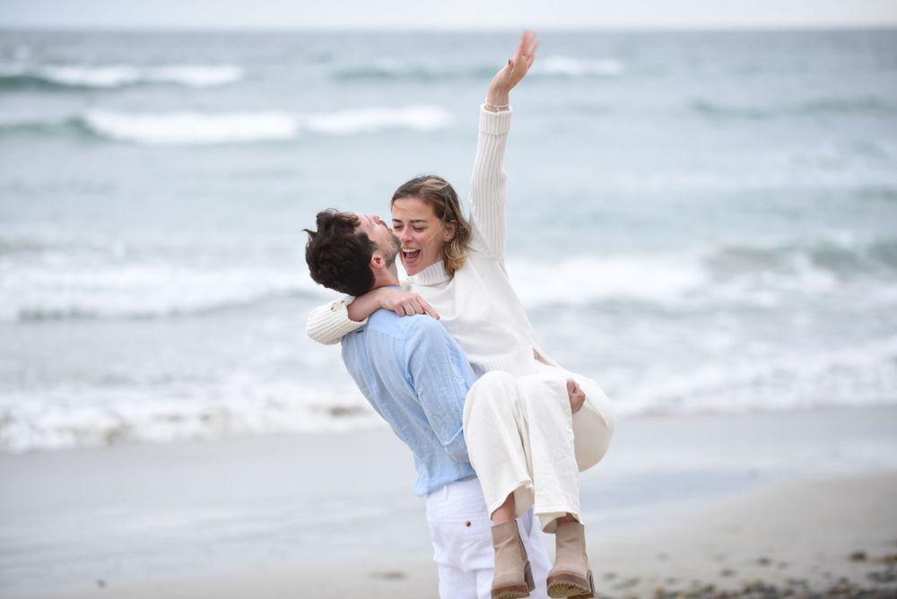 Two people dancing and embracing joyfully on a scenic beach with waves crashing behind them.