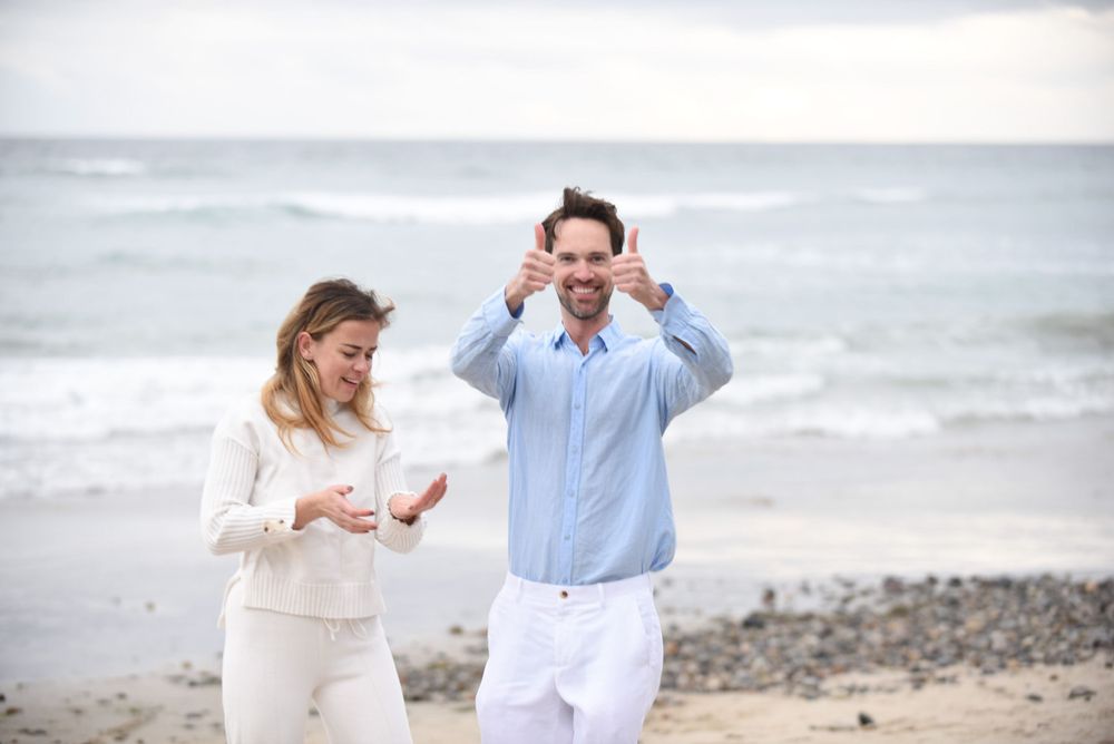 A couple in white and blue outfits dancing playfully on a coastal beach.