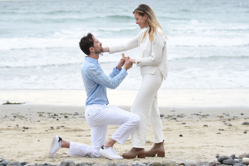 A surprise proposal moment captured on a scenic beach with crashing waves in the background.