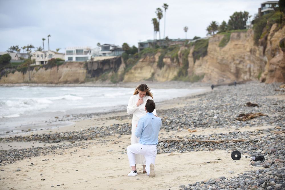 Two people walking along a rocky beach with coastal cliffs and ocean waves in the background.