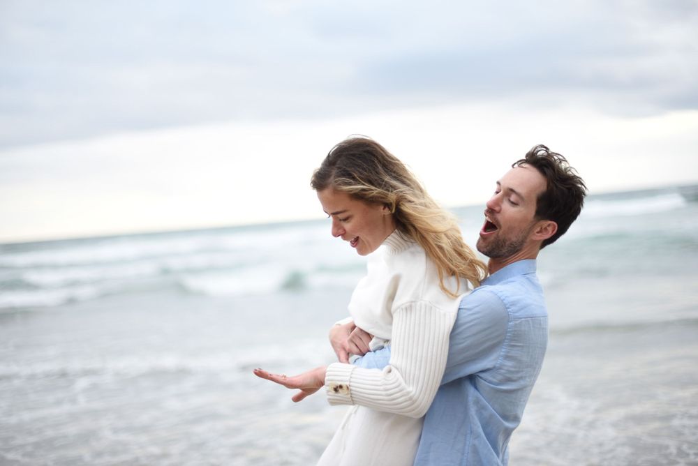 A tender moment between a couple in white and blue clothing on an overcast beach.