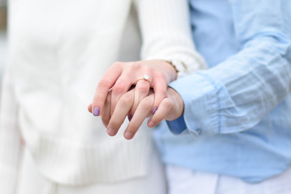 Close-up of intertwined hands showing an engagement ring against white and blue clothing.