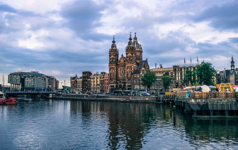 Historic Saint Nicholas Church reflects in Amsterdam harbor waters under dramatic sky.