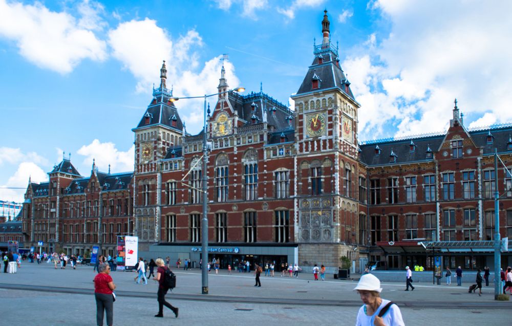 Amsterdam Central Station's grand Victorian facade on a sunny day with tourists in the square.