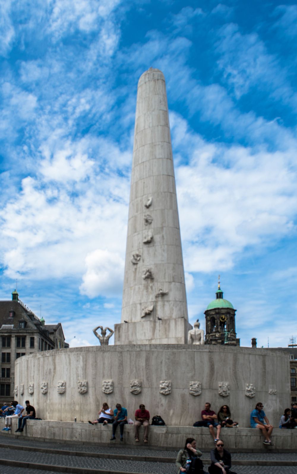 Tall white monument obelisk against blue sky with people sitting around its base in Amsterdam.