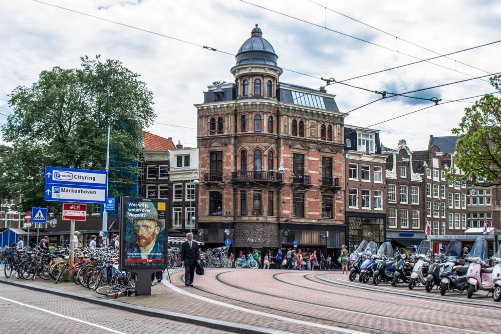 Distinctive domed historic building on an Amsterdam corner with tram lines and parked scooters.