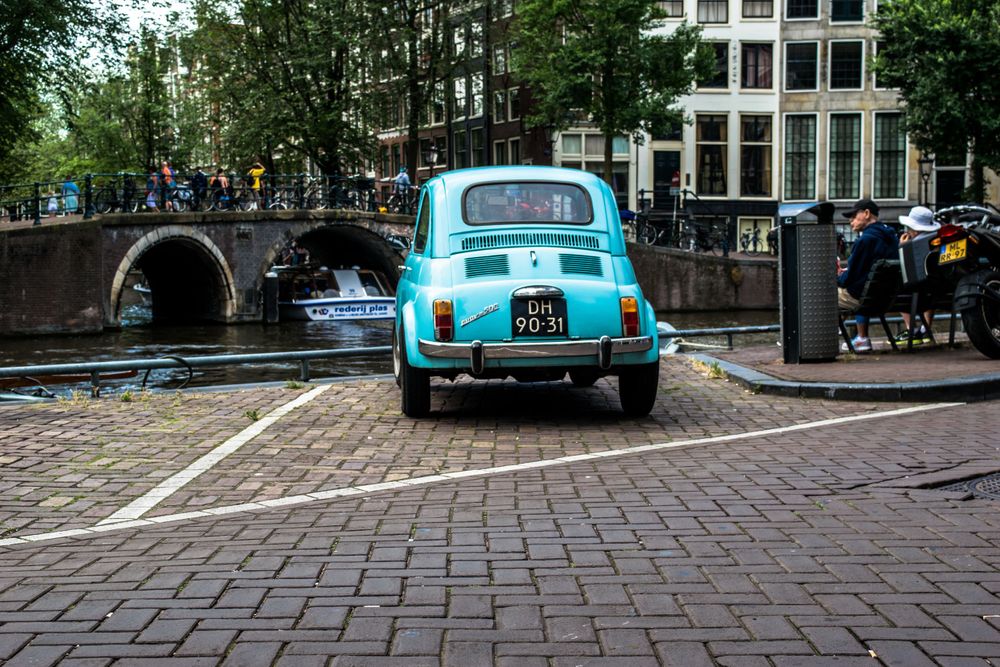 Vintage turquoise Fiat 500 parked on cobblestone street by Amsterdam canal bridge.