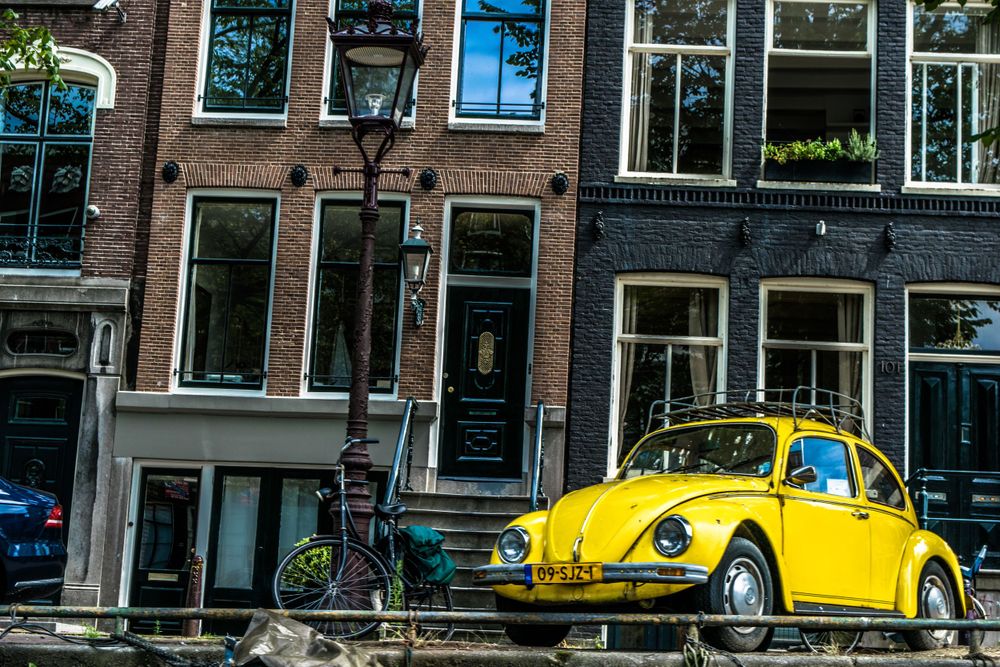 Yellow vintage Volkswagen Beetle parked along Amsterdam canal with traditional architecture.