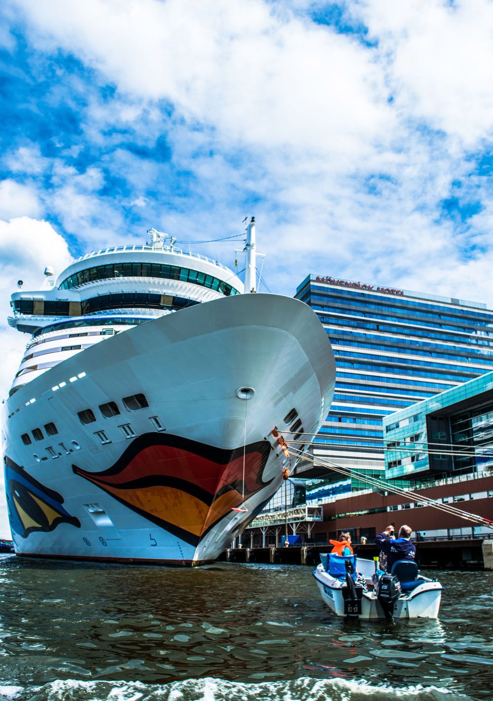 Large cruise ship with distinctive bow art docked at port while small boat passes in foreground.