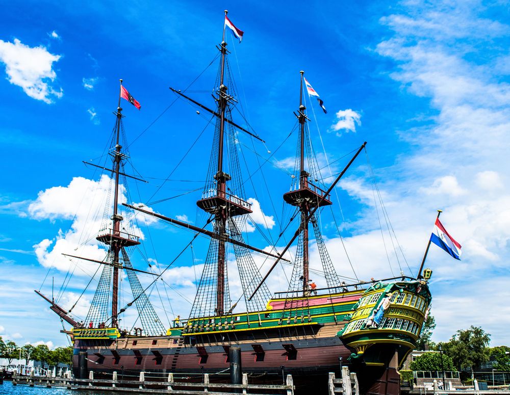 Historic tall ship with three masts and green hull displayed against bright blue sky with white clouds.