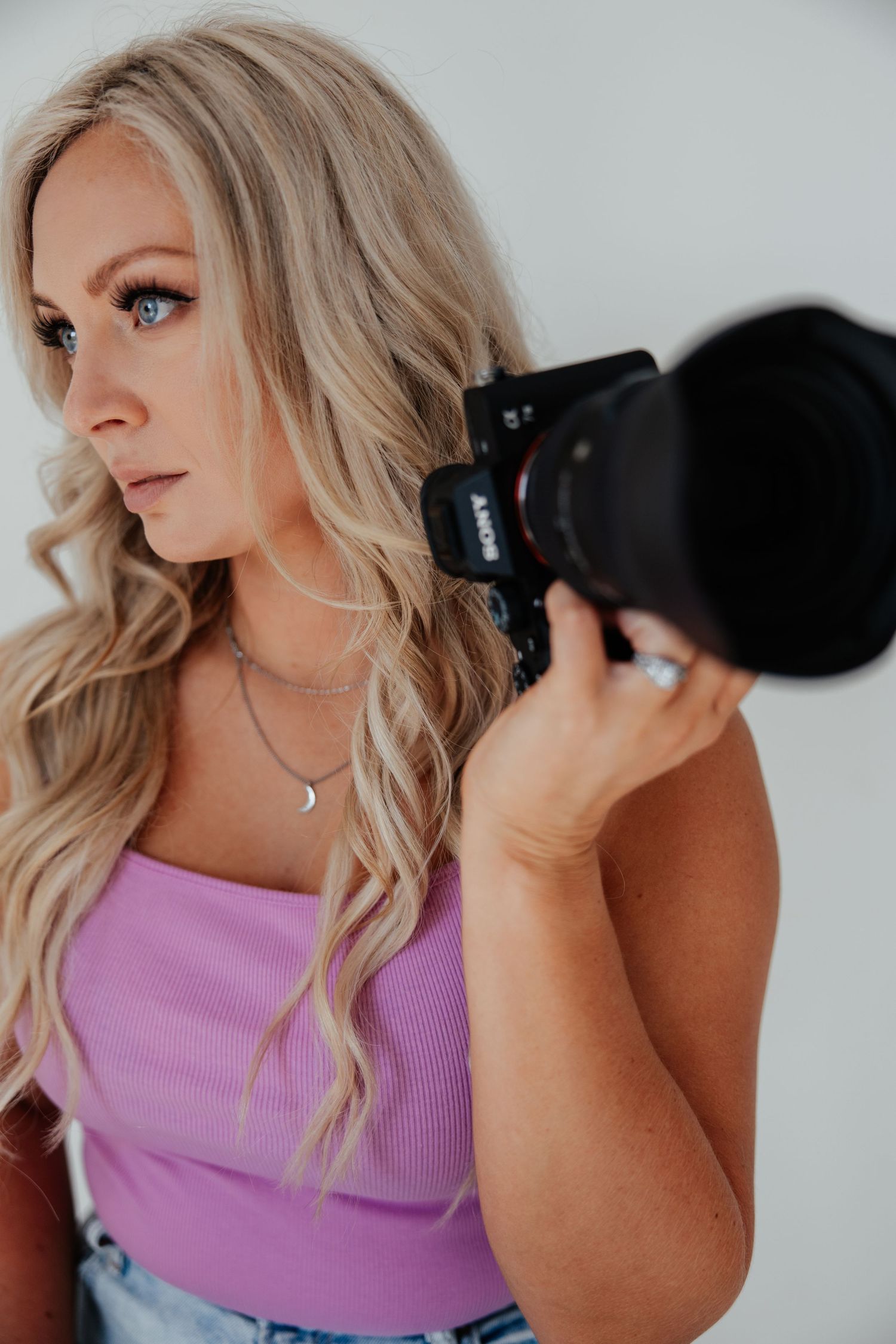 A professional photographer in a pink tank top holds a camera with a long lens while posing against a grey background.