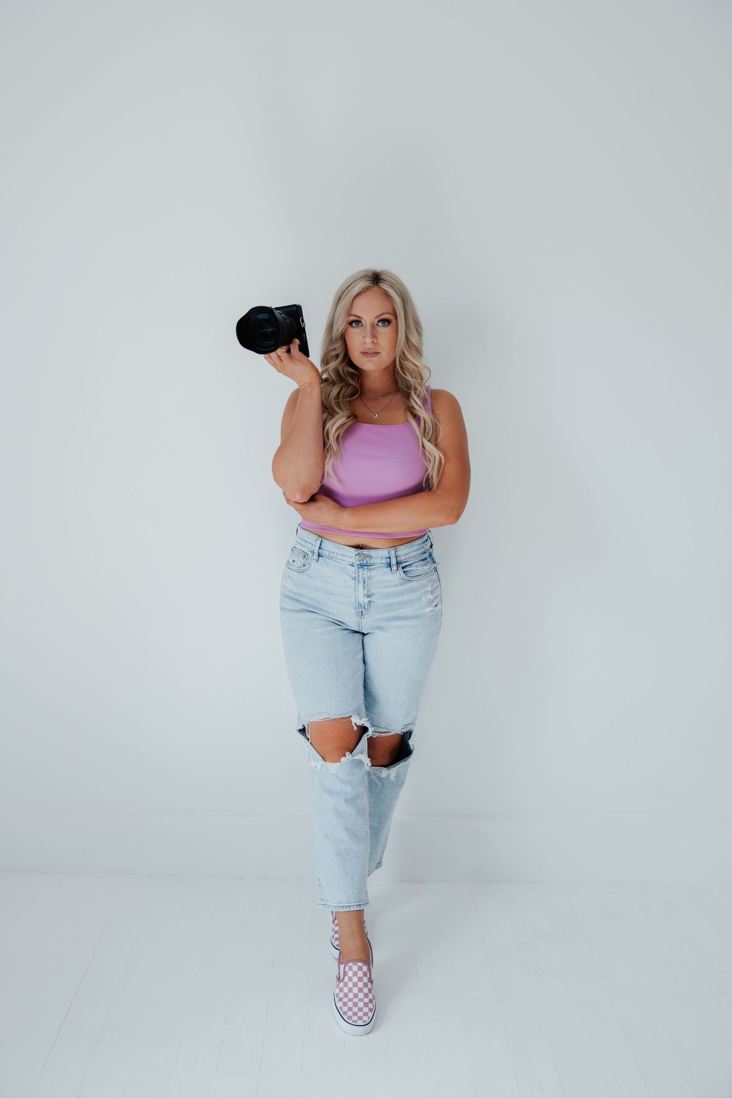 A photographer in distressed jeans and pink top poses with camera against white backdrop.