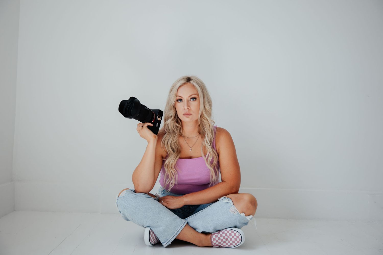 A photographer sits cross-legged on white floor holding professional camera equipment.