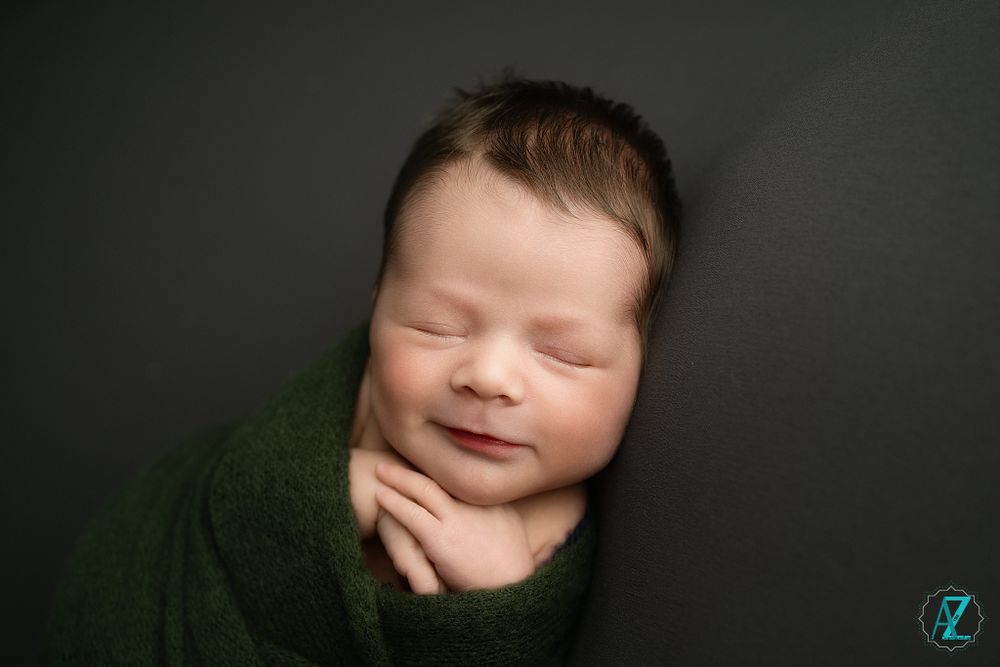 Newborn portrait of a sleeping baby wrapped in green against a dark background.
