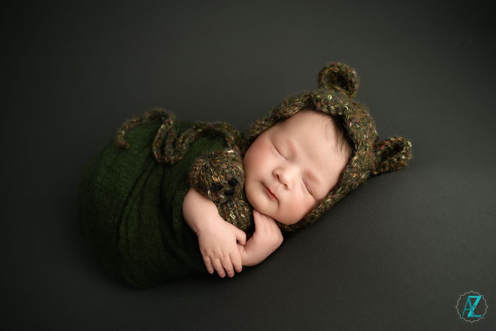 Sleeping newborn wearing a brown bear bonnet and wrapped in green fabric.
