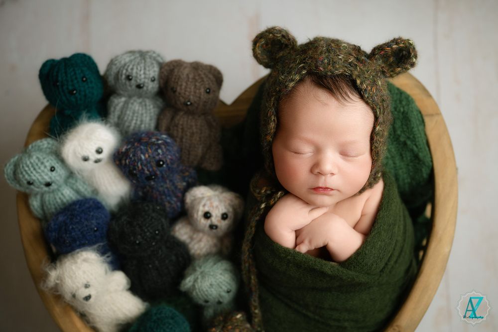 Sleeping newborn in green wrap posed in wooden heart bowl with plush animals.