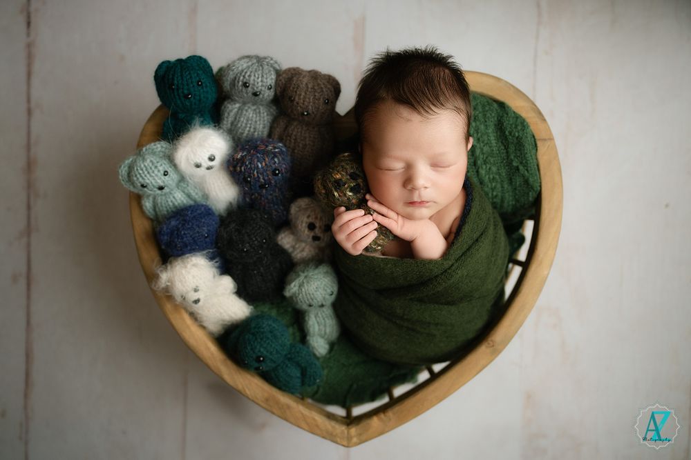 Newborn baby sleeping in heart-shaped basket surrounded by stuffed toys.