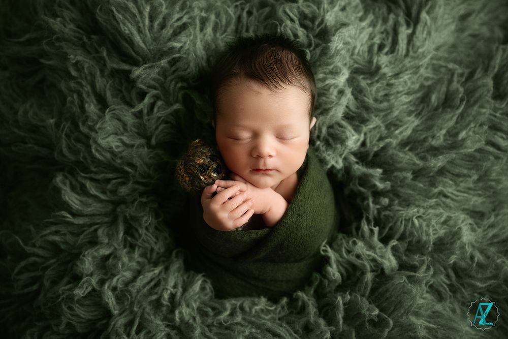 Sleeping newborn wrapped in green on textured dark green fur background.