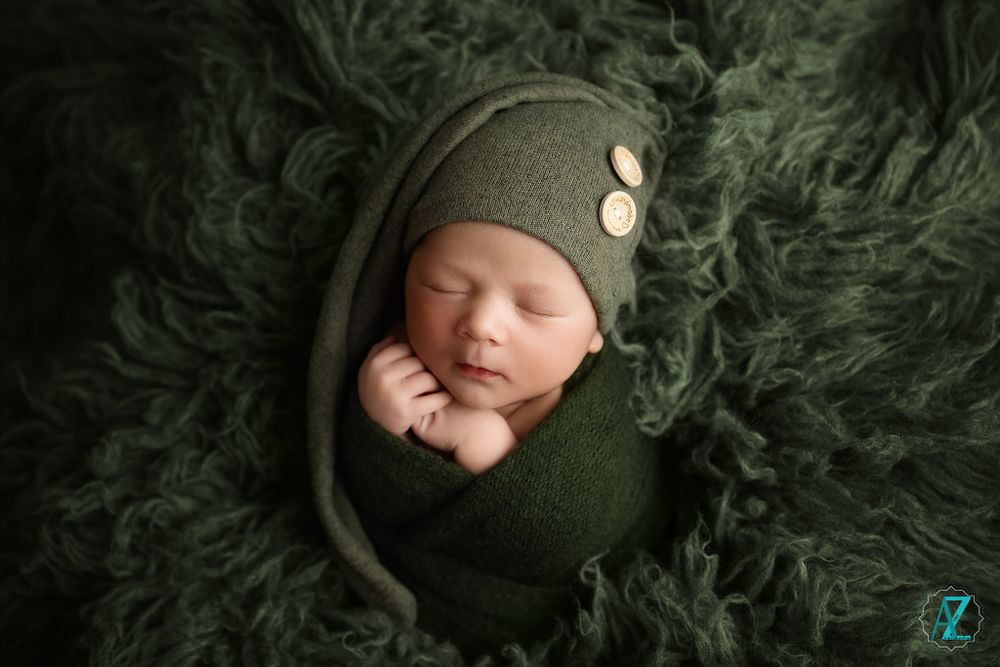 Peaceful sleeping newborn wrapped in green wearing a knit cap on furry backdrop.