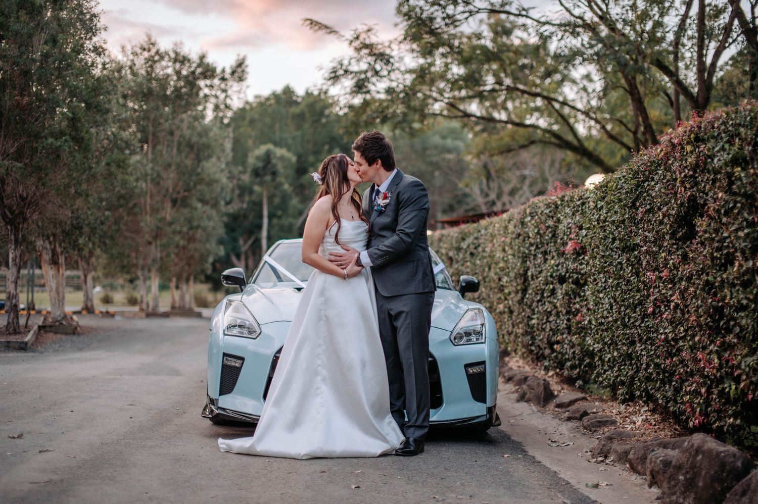 Wedding couple poses against white car on tree-lined road during sunset.