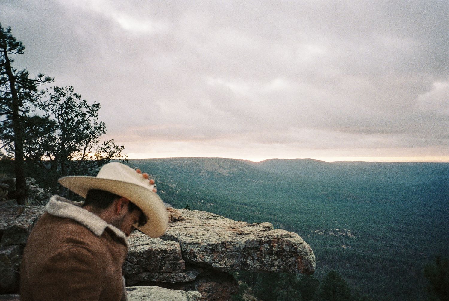 A person in a cowboy hat gazes out over a misty mountain landscape at sunset.