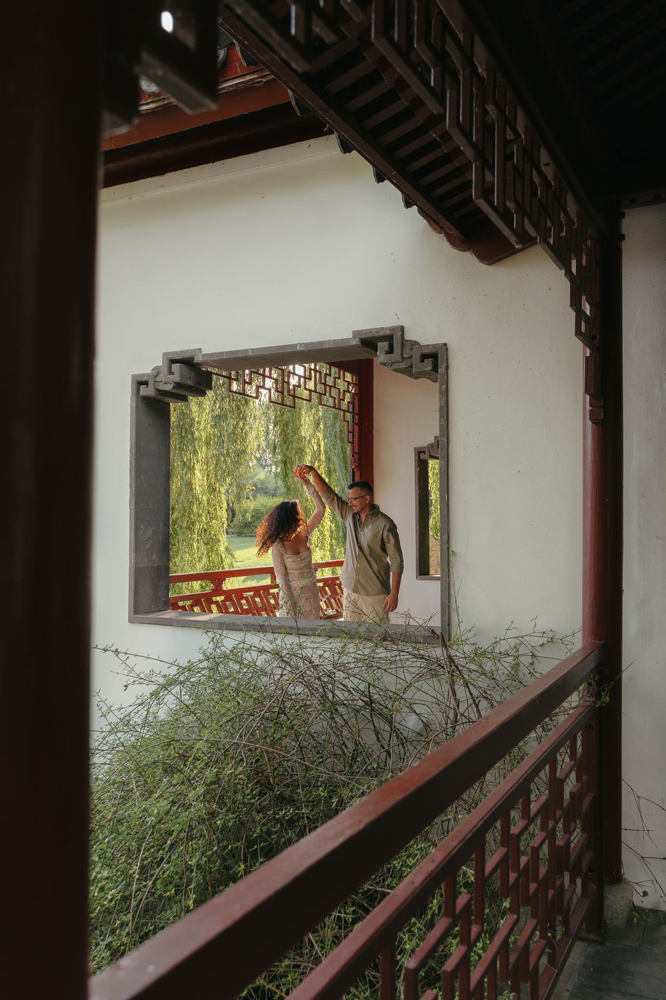 View through decorative wooden window frame of couple with willow trees beyond.