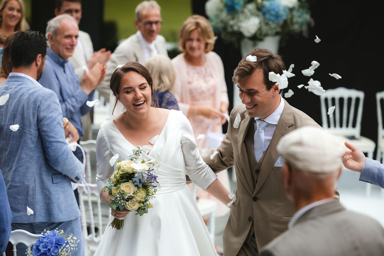 A joyful wedding celebration with guests throwing white confetti as newlyweds walk together outdoors.