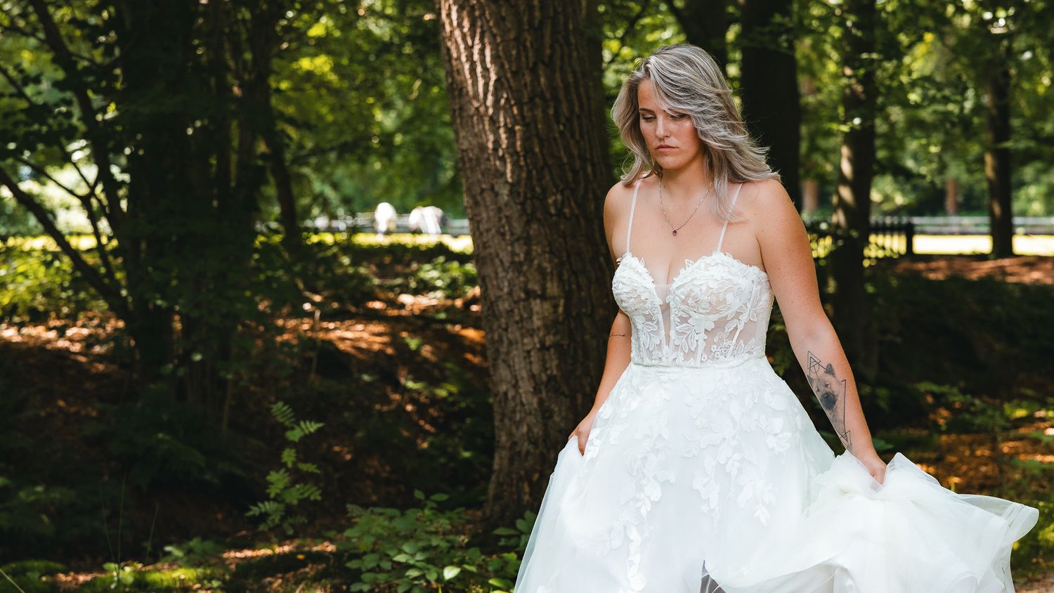A bride in a white lace wedding dress stands gracefully among autumn trees in a garden setting.