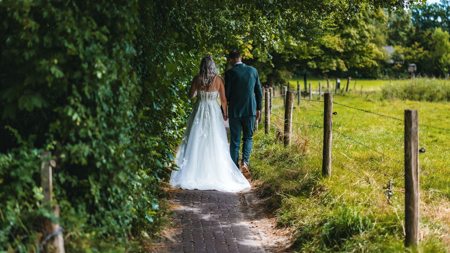 A couple walking down a rural path surrounded by green foliage and wooden fence posts on a sunny day.