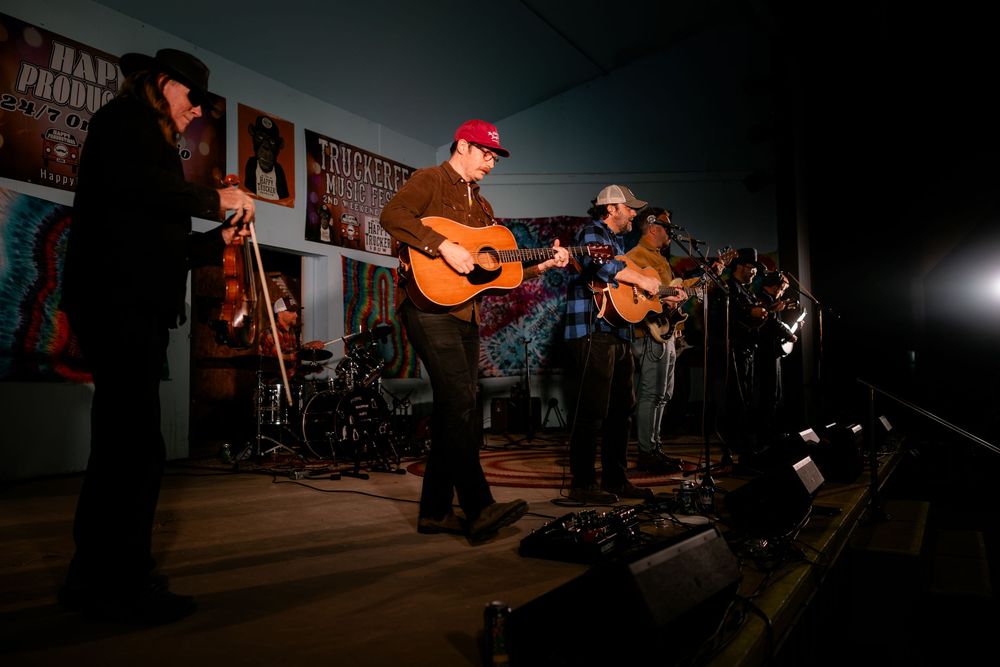 Musicians perform on stage at a dimly lit venue with acoustic instruments and microphones.