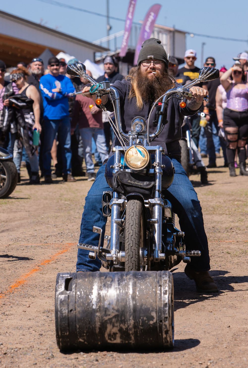 A bearded motorcyclist riding a custom bike with a large barrel attachment at a motorcycle event with spectators.