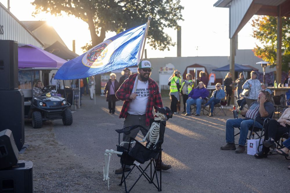 People gather at an outdoor event with tents and vehicles visible in the evening sunlight.