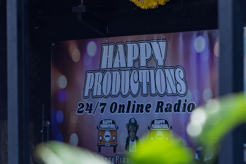 A radio station banner with colorful text and lighting displays 'Happy Productions 24/7 Online Radio' against a dark background.