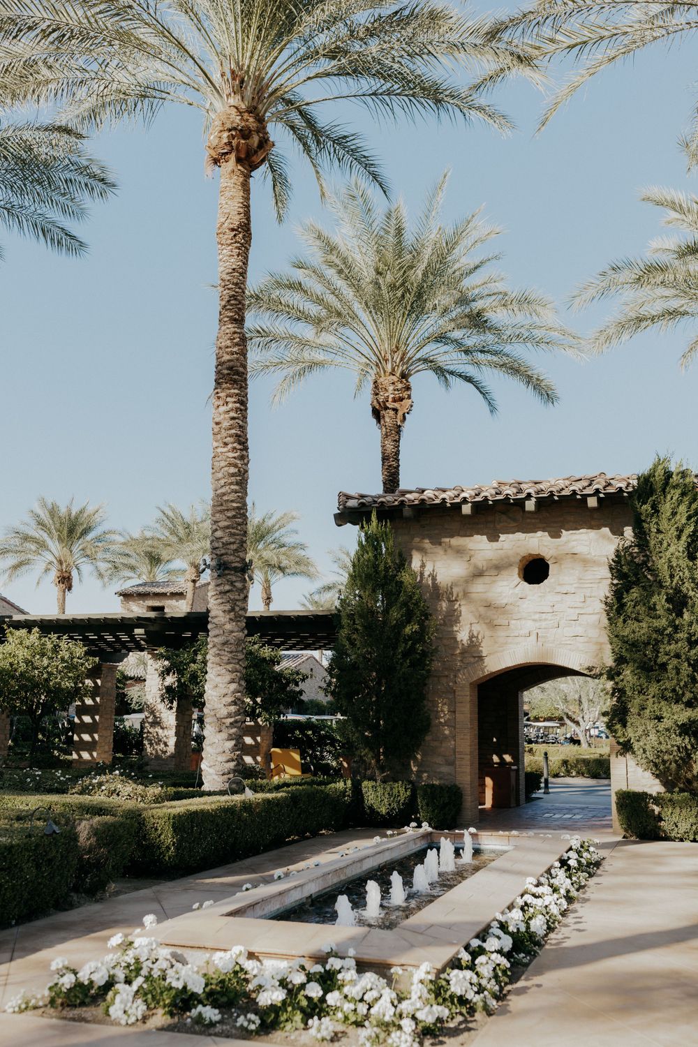 Elegant garden pathway with palm trees and white flowers leading to a stone archway.