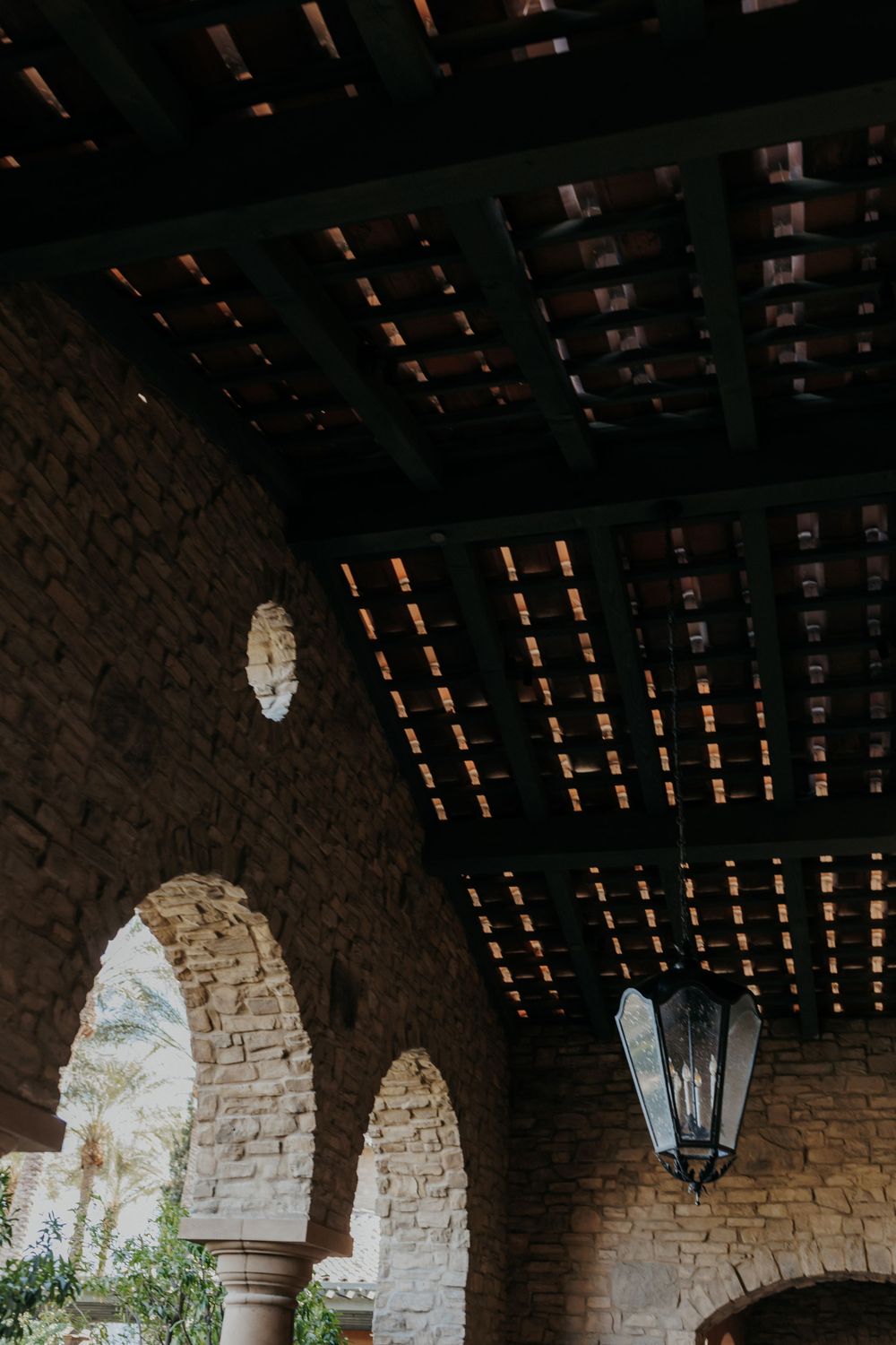 Rustic stone archway with wooden beam ceiling and hanging lantern in Spanish colonial style.