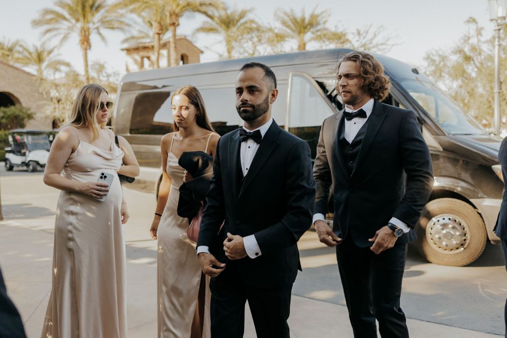 Wedding party in formal attire gathered outside a luxury vehicle with palm trees in the background.